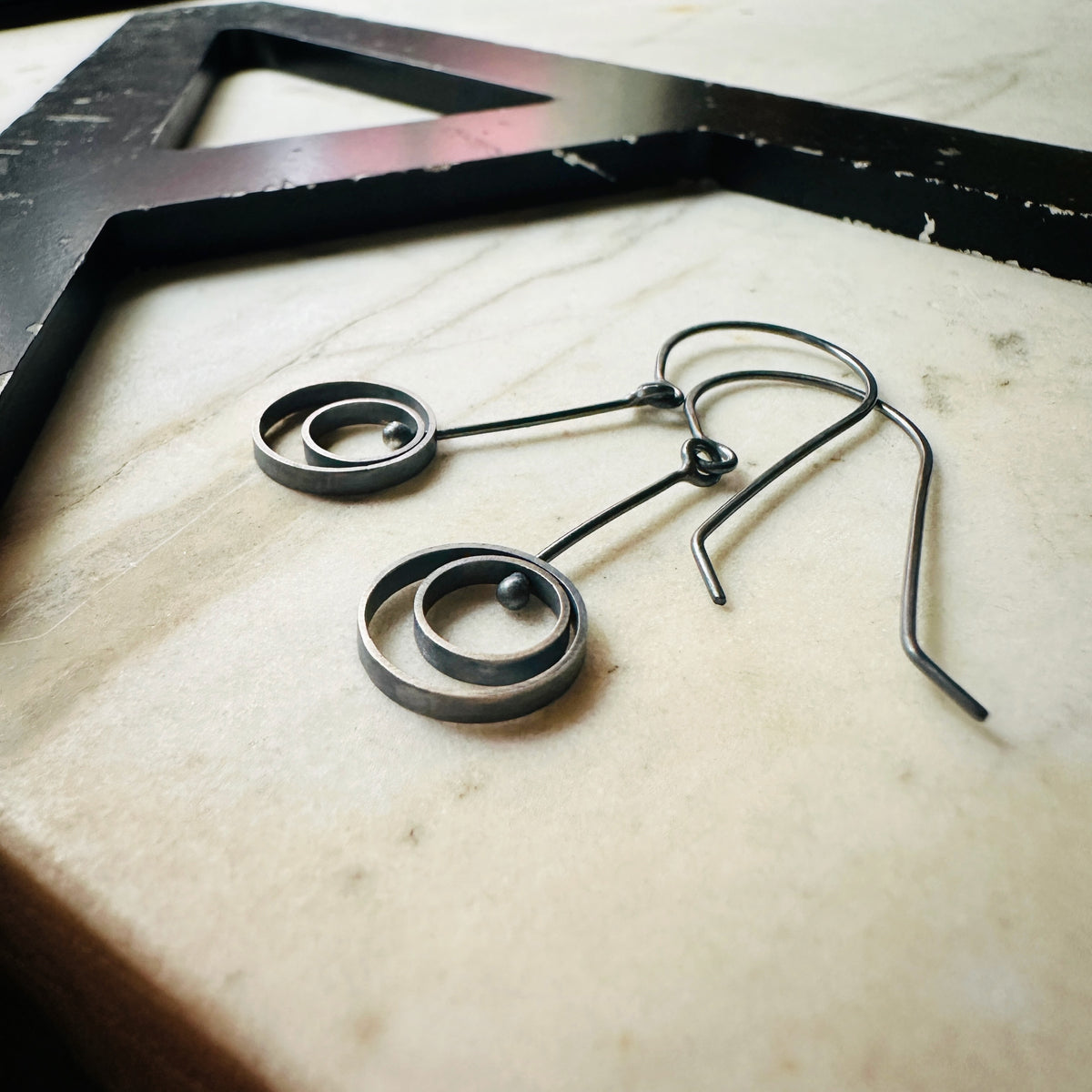 A pair of oxidized sterling silver drop earrings with circular spiral designs, resting on a light beige marble surface next to a black wooden frame.