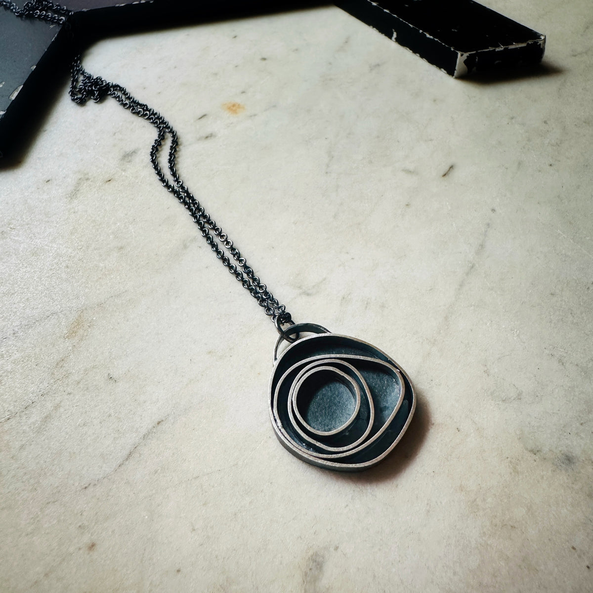 An oxidized sterling silver concentric circle pendant necklace on an oxidized silver chain, resting on a light beige stone surface.