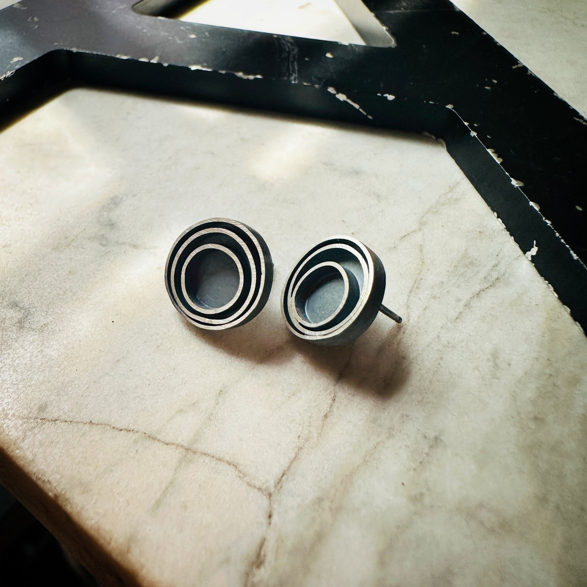 A pair of round stud earrings with a concentric circular design in a oxidized sterling silver, resting on a light beige marble surface.