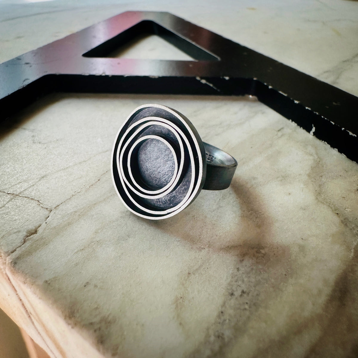 oxidized silver ring with concentric circle designs, with a large black metal letter 'A' in the background.