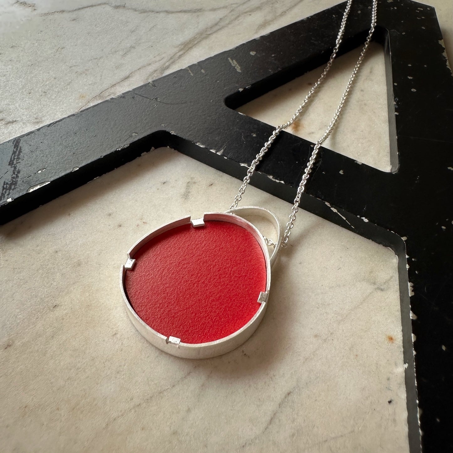 A silver pendant necklace with a circular bezel holding a vibrant red Formica textured insert, resting on a black decorative letter 'A' over a light marble surface.