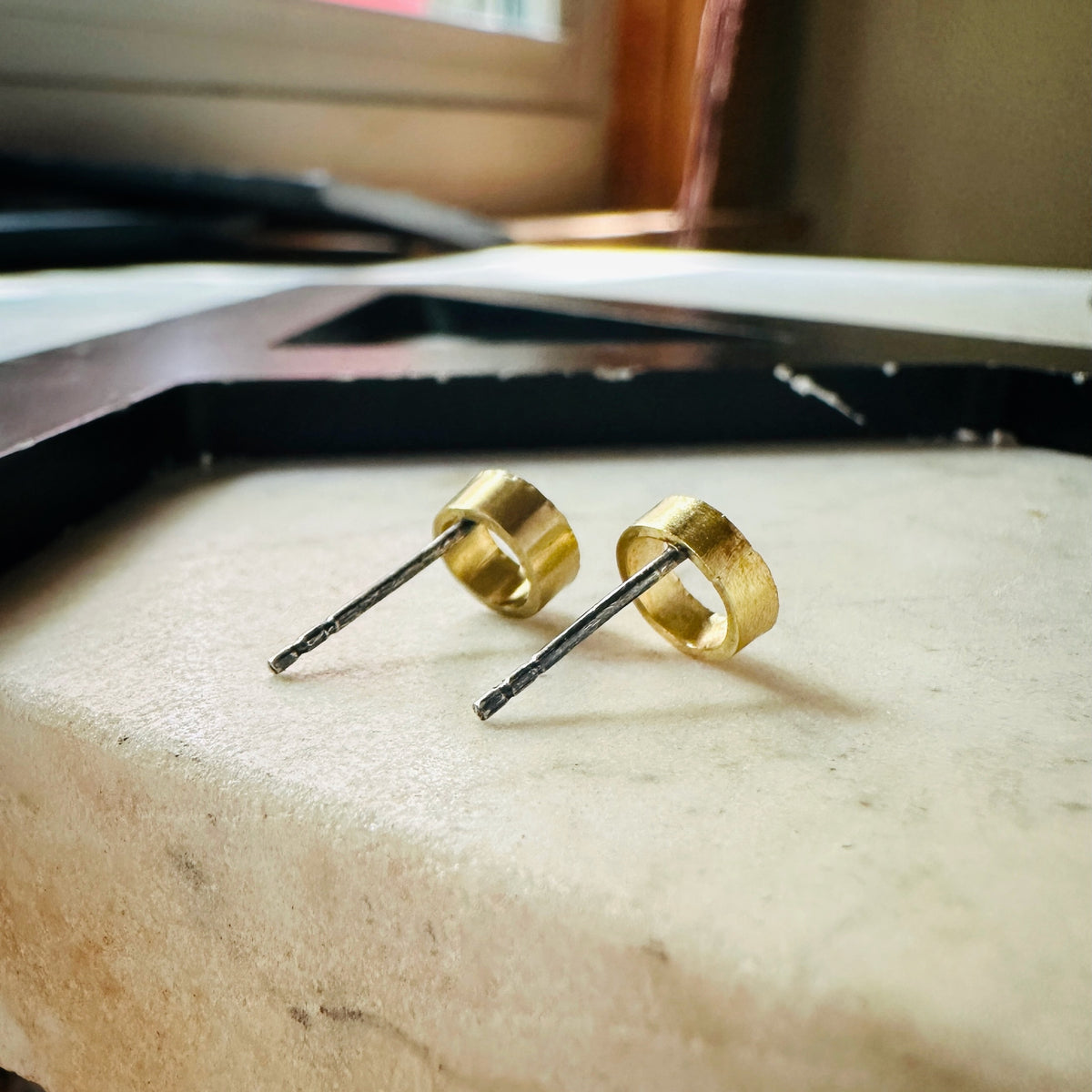 A pair of gold hoop stud earrings with dark metal posts, resting on a light beige stone surface.