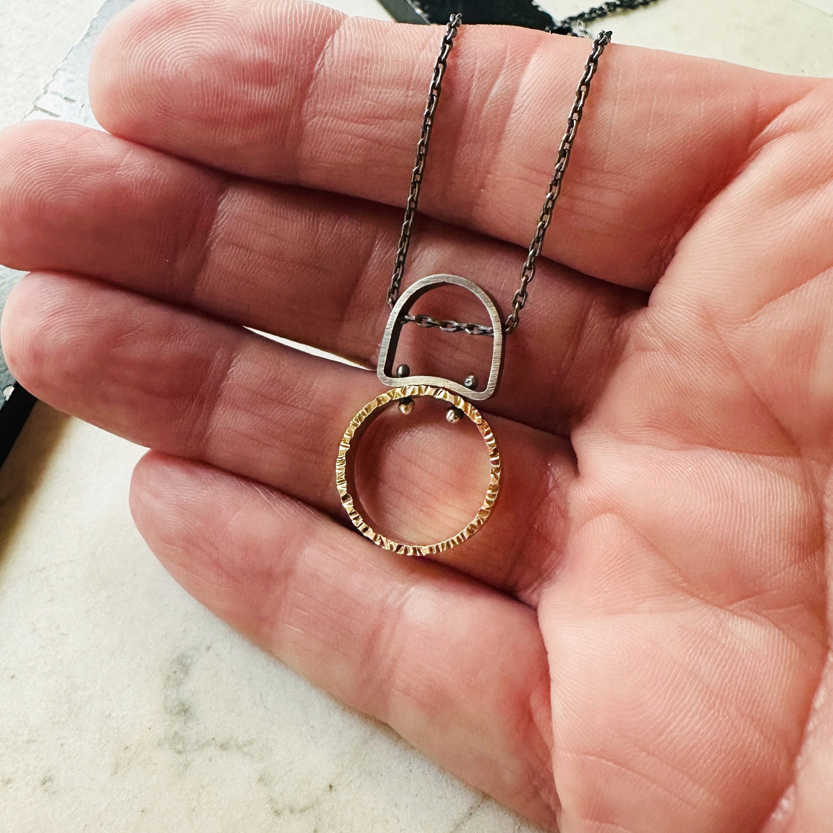 a hammered gold circle pendant with a oxidized silver chain and bail on a hand
