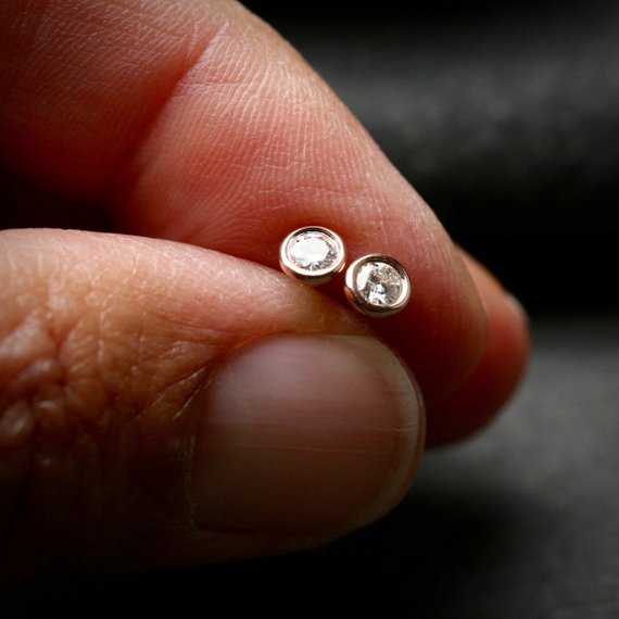 14k rose gold 3mm Moissanite stud earrings. They are bezel set and shown being held in two fingers for scale.