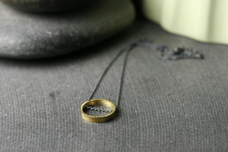 A half inch circle of 18k yellow gold with a hammered texture. It is on an oxidized silver chain that runs through the circle.