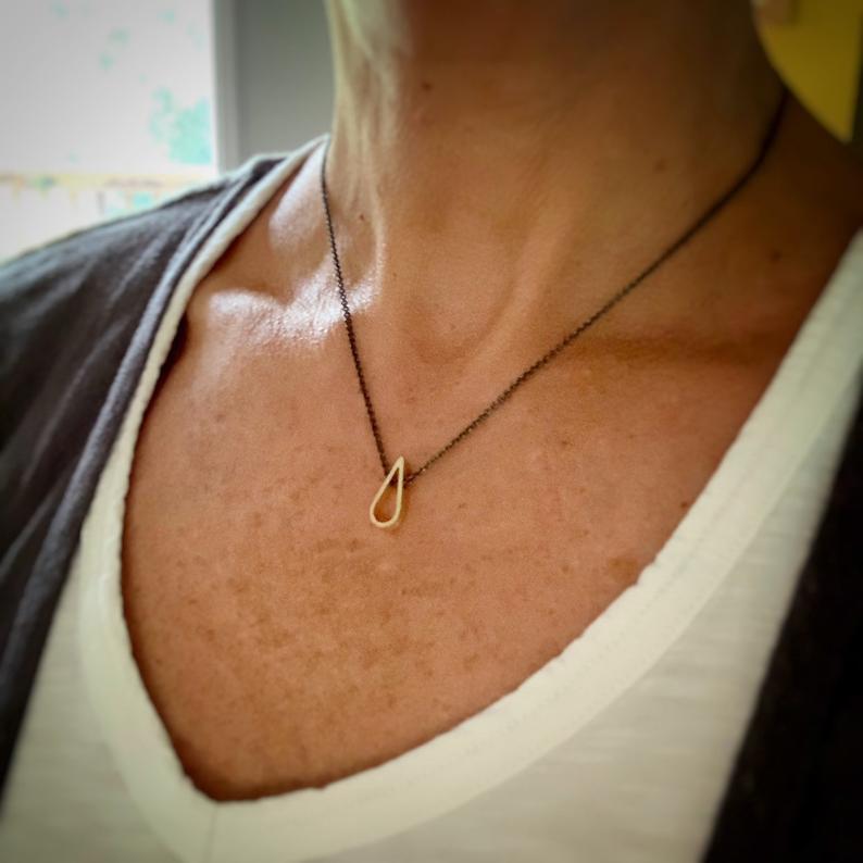 tiny 18k yellow gold teardrop pendant with a darkened silver chain being worn by a white woman with a white t shirt and dark sweater.