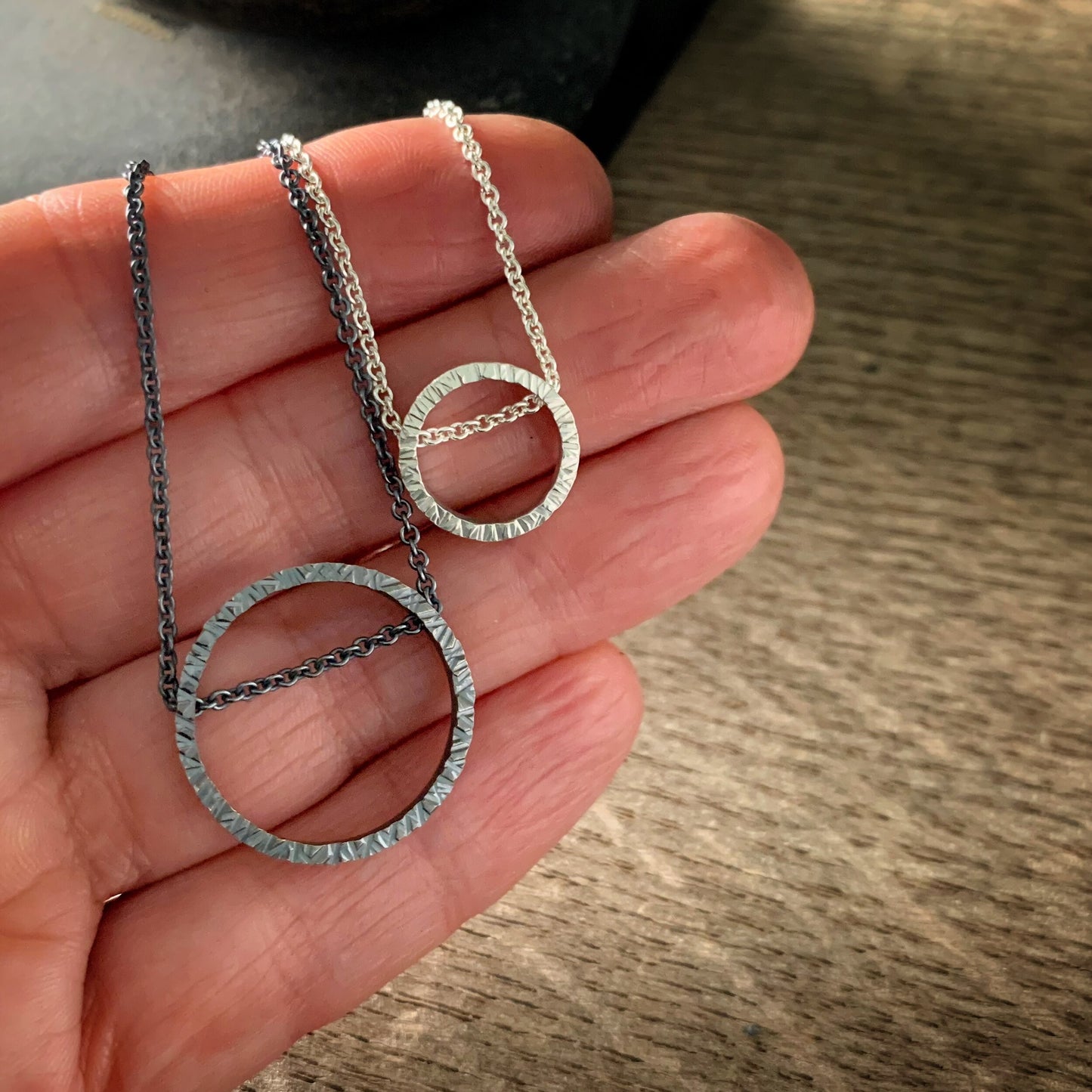 two pendants, one large and one small, in sterling silver. They are circles with a hammered texture and the chain runs through the circle. One is darkened silver and the other bright silver. Shown on a hand for scale