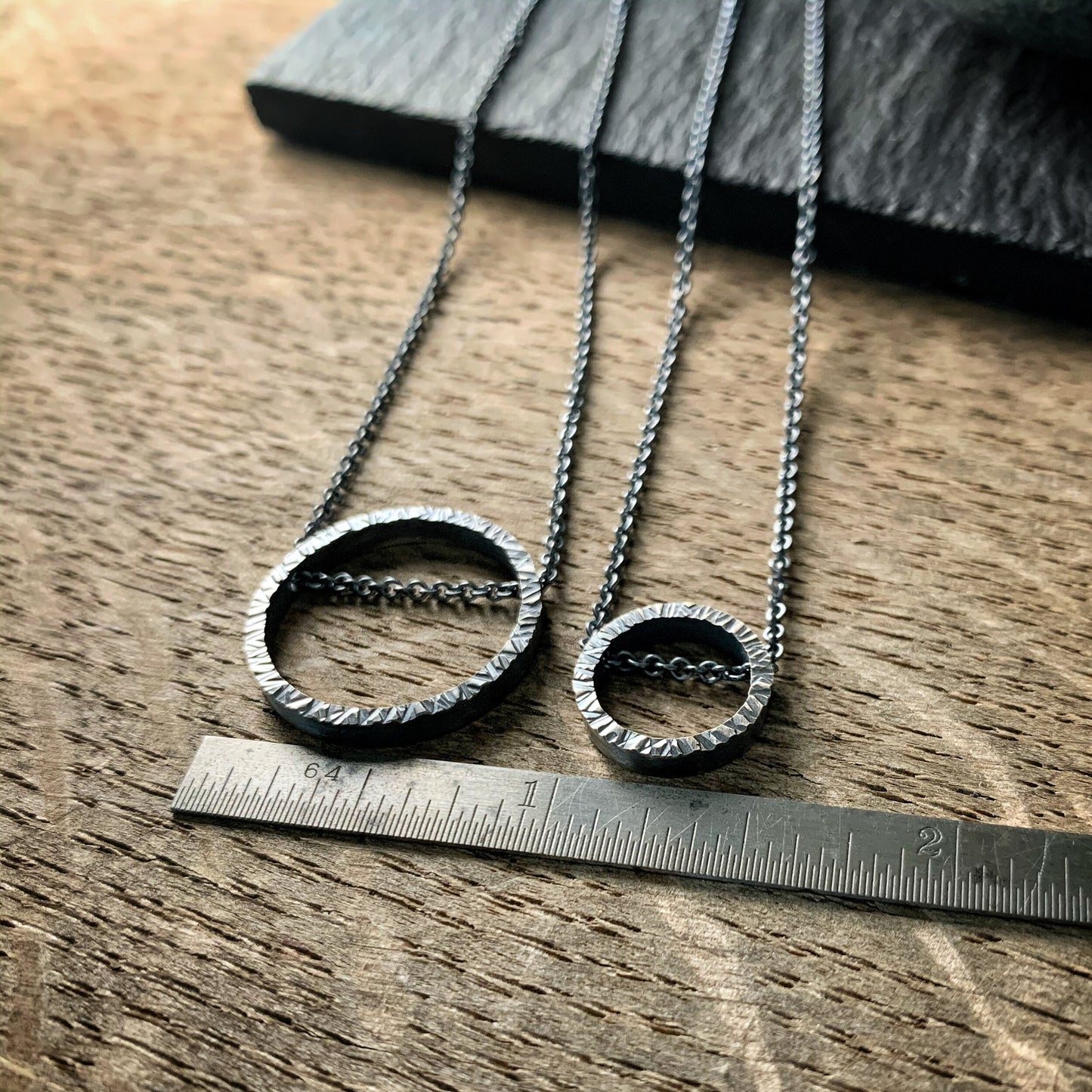 two pendants, one large and one small, in sterling silver. They are circles with a hammered texture and the chain runs through the circle. One is darkened silver and the other bright silver. Shown on a ruler for scale.