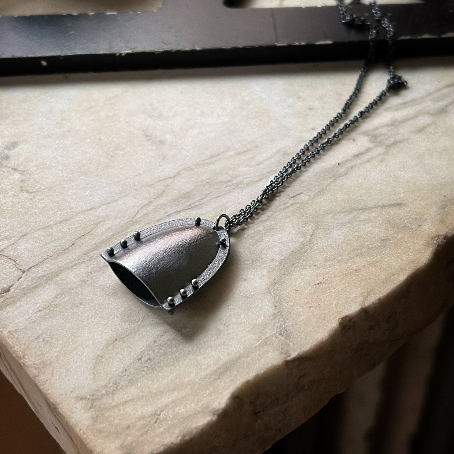 a bell shaped pendant with a modern industrial look. It's darkened silver with pin rivets and a chain