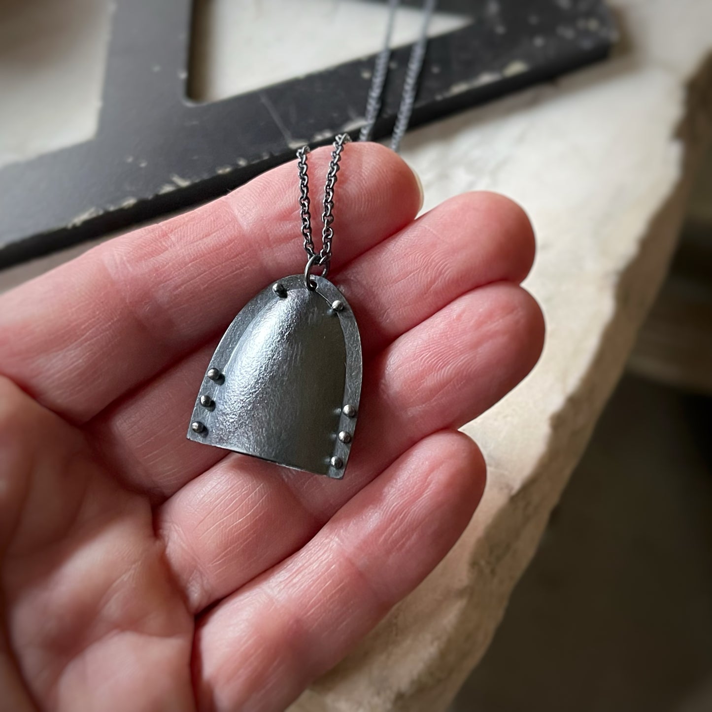 a bell shaped pendant in oxidized silver with tiny pin rivets shown in a hand for scale