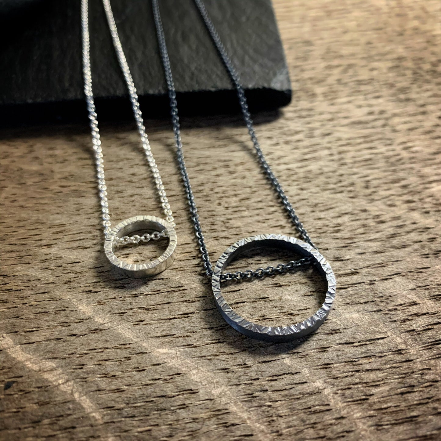 two pendants, one large and one small, in sterling silver. They are circles with a hammered texture and the chain runs through the circle. One is darkened silver and the other bright silver