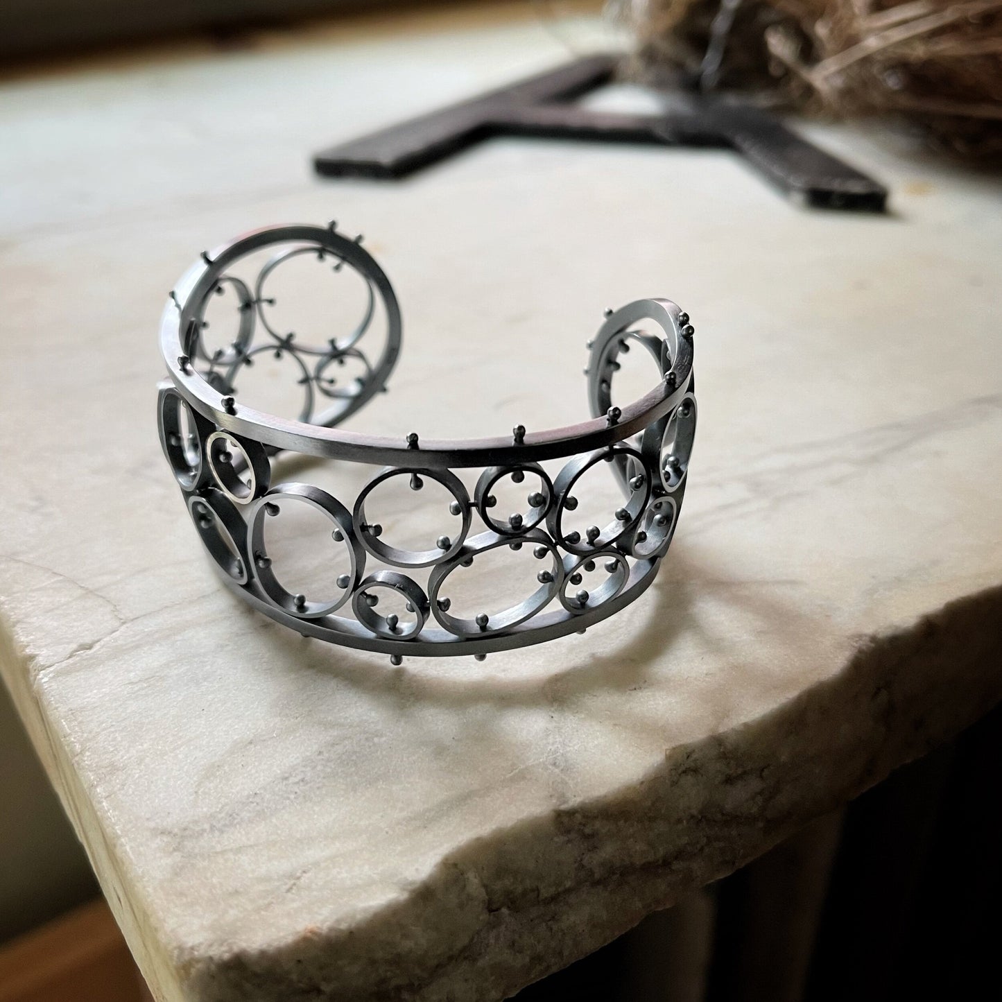 A sterling silver bracelet with a brushed finish and oxidized details, featuring a series of linked metal cells, displayed on a textured surface.
