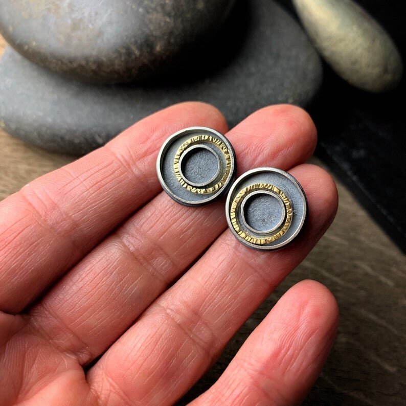 oxidized silver earrings with concentric circles, one of each is yellow gold. Shown on a hand for scale
