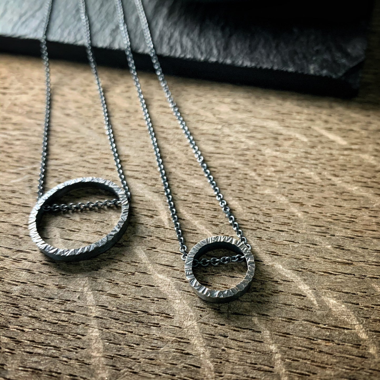 two pendants, one large and one small, in sterling silver. They are circles with a hammered texture and the chain runs through the circle. Both are oxidized silver.