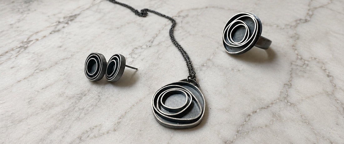three oxidized sterling silver pieces of jewelry; a pair of earrings, a pendant and a ring.  They have concentric circles and are lying on marble
