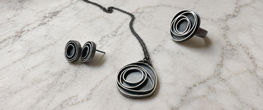 three oxidized sterling silver pieces of jewelry; a pair of earrings, a pendant and a ring.  They have concentric circles and are lying on marble