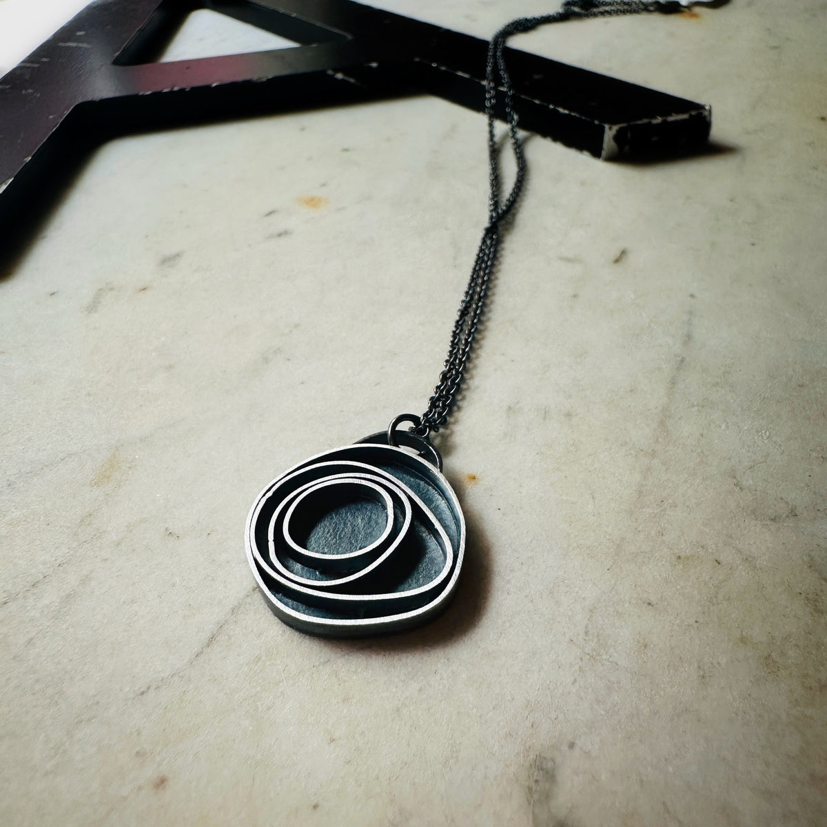 An oxidized sterling silver concentric circle pendant necklace on an oxidized silver chain, resting on a light beige stone surface.