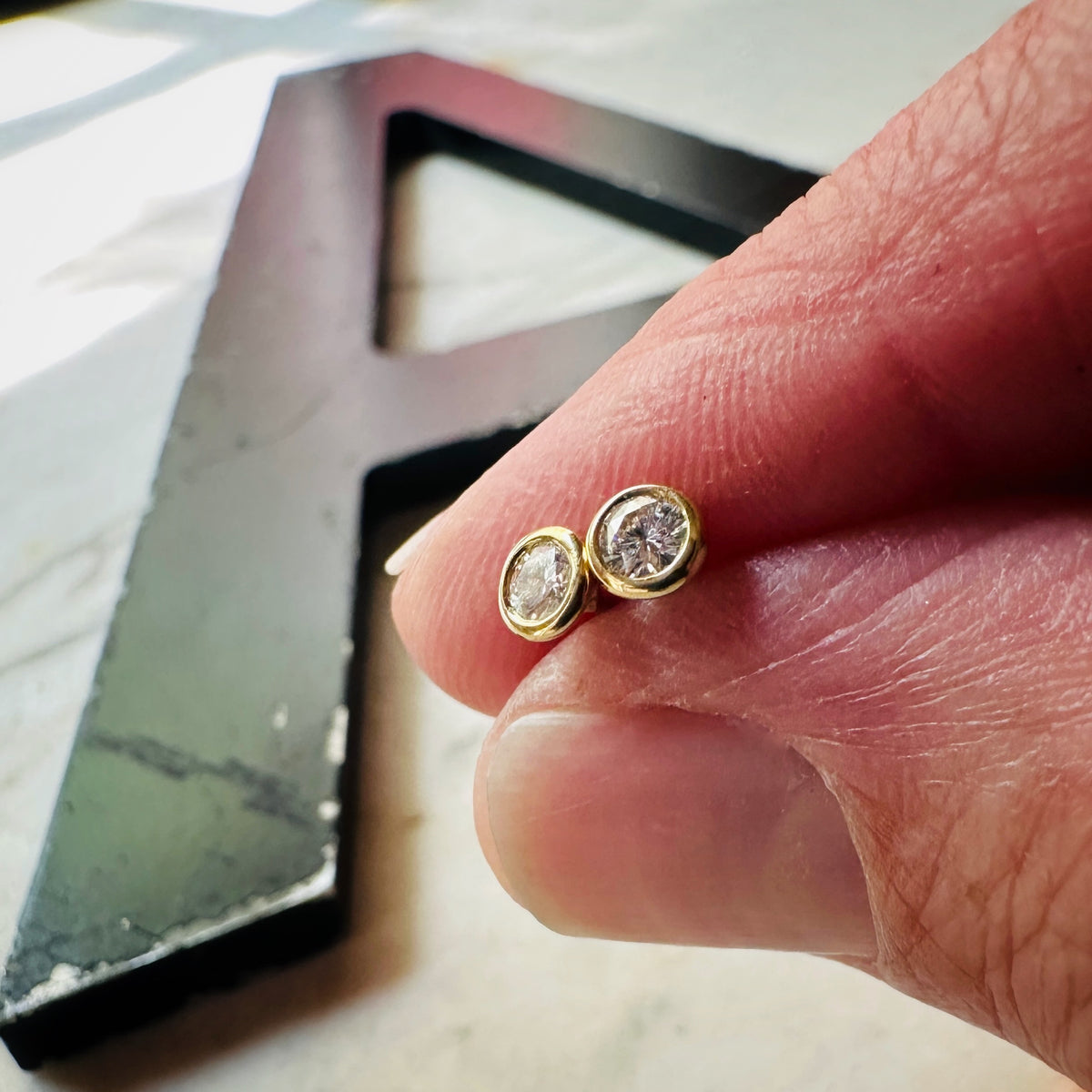 18k yellow gold 3mm Moissanite bezel set stud earrings on a marble background shown being held in fingers for scale