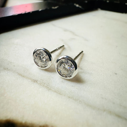 A pair of 5mm Moissanite bezel-set stud earrings sterling silver, resting on a light textured surface.