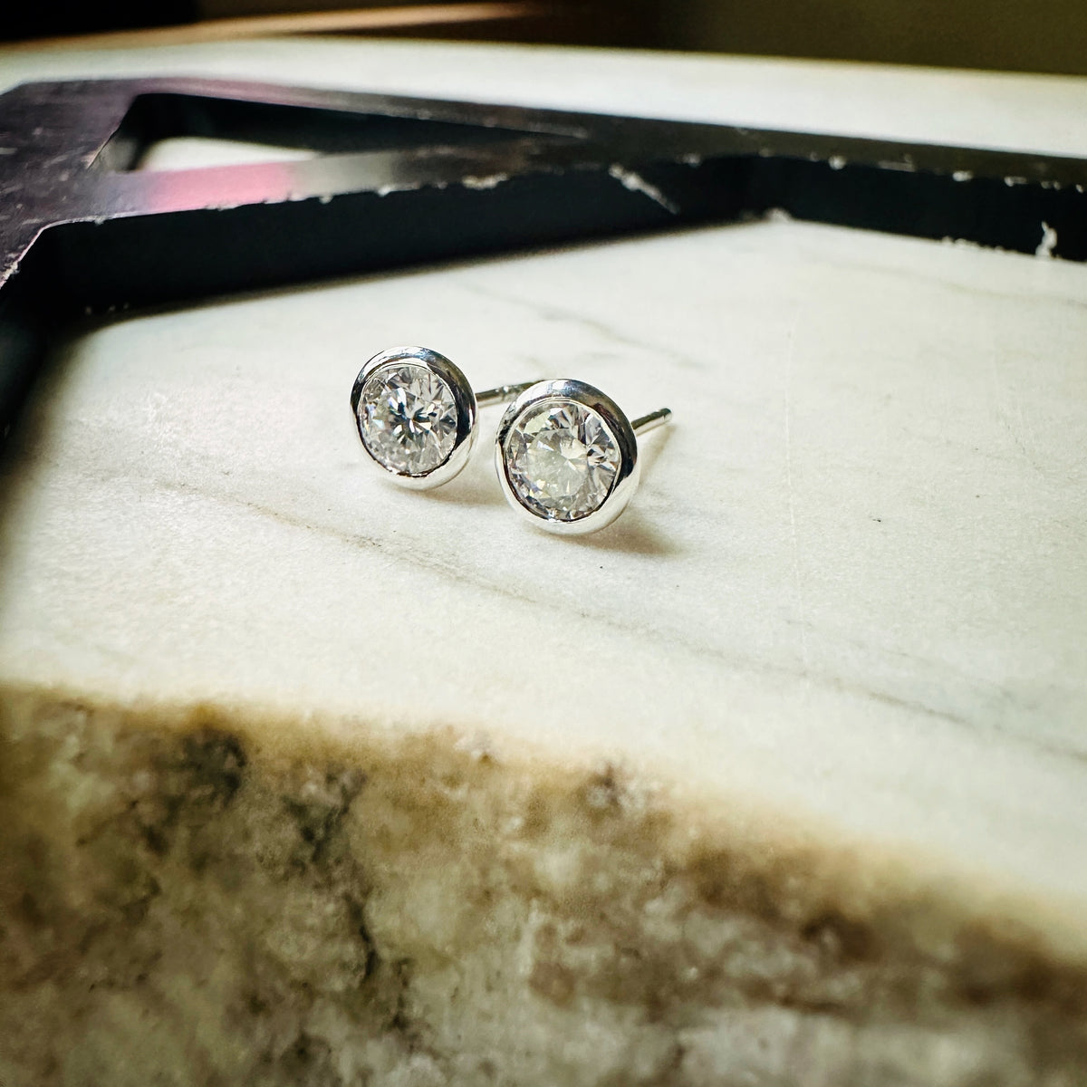 A pair of 5mm Moissanite bezel-set stud earrings sterling silver, resting on a light textured surface.