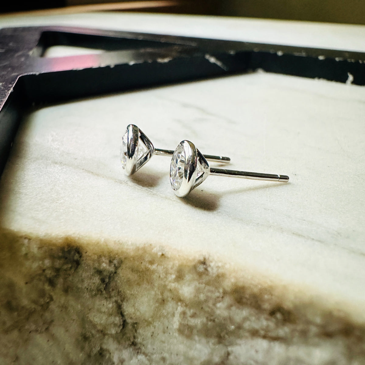 A pair of 5mm Moissanite bezel-set stud earrings sterling silver, resting on a light textured surface seen from the side.