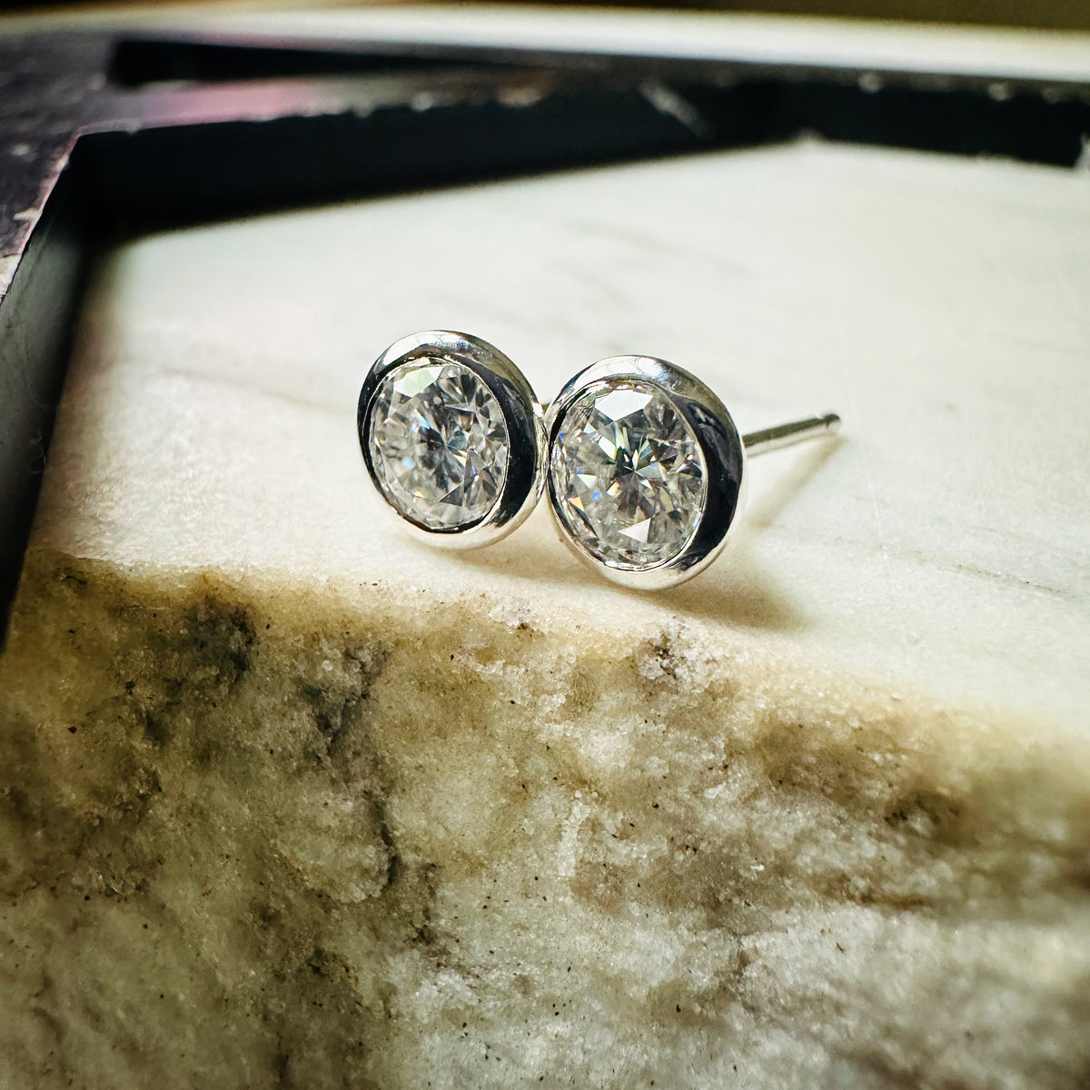 A pair of 5mm Moissanite bezel-set stud earrings sterling silver, resting on a light textured surface.