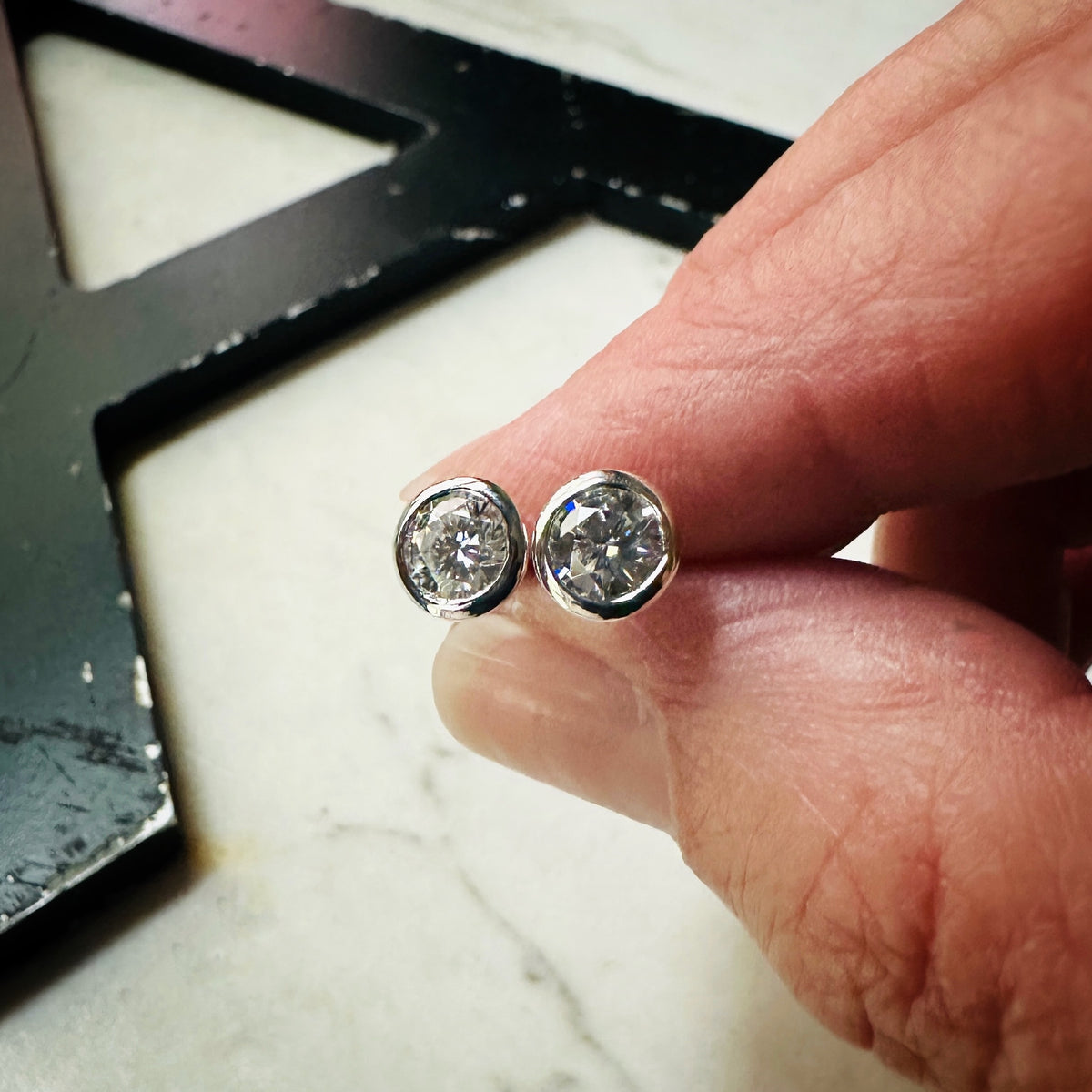 A pair of 5mm Moissanite bezel-set stud earrings sterling silver, resting on a light textured surface being held in a hand for scale