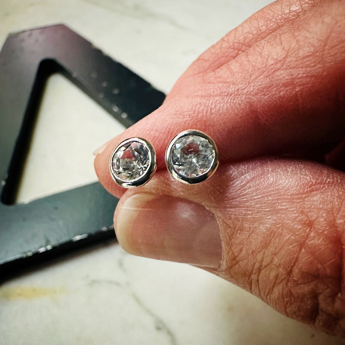 5mm white topaz bezel set stud earrings in sterling silver held in fingers to show scale 