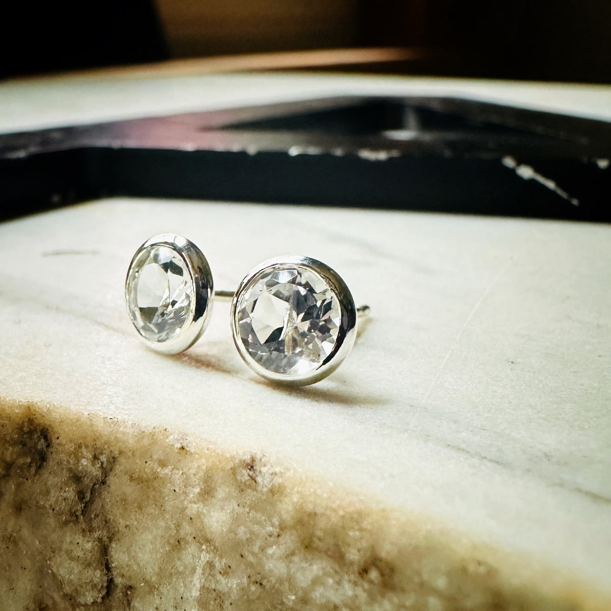 A pair of 6mm white topaz round bezel-set stud earrings in silver bezel settings, resting on a light stone surface.