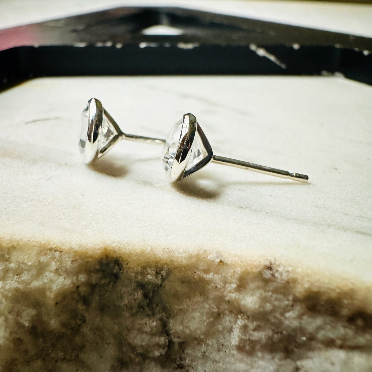 A pair of 6mm white topaz silver stud earrings shown from the side, resting on a textured light beige stone surface.