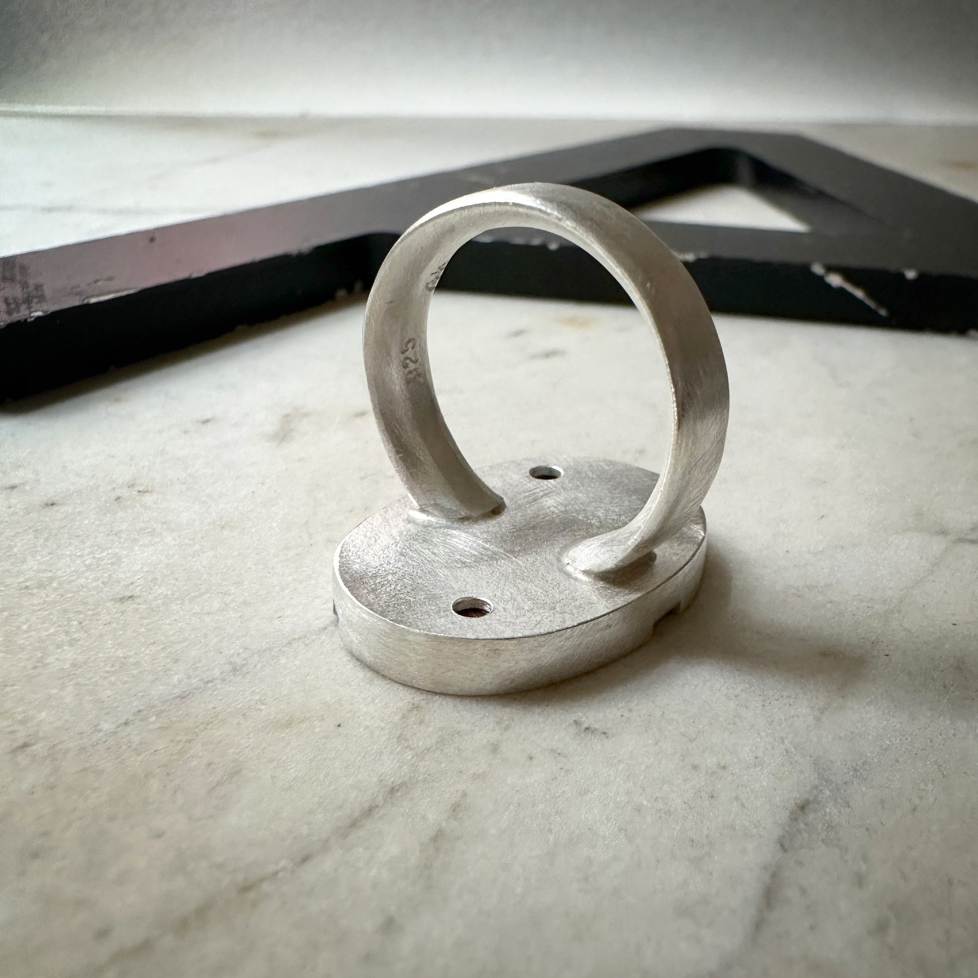 A handmade silver ring with a thick, rounded band and a flat, circular base featuring two pre-drilled holes, displayed on a light marble surface.