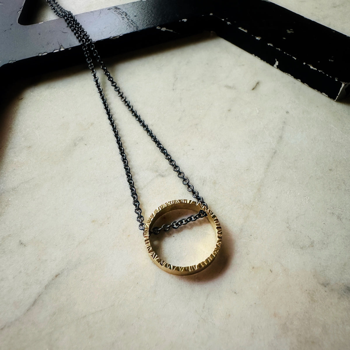 yellow gold hammered texture circle pendant with oxidized sterling silver chain on a marble surface 