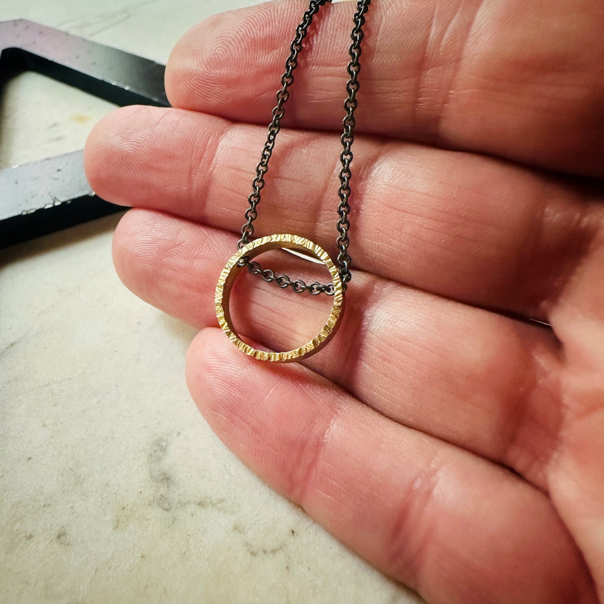 yellow gold hammered texture circle pendant with oxidized sterling silver chain on a marble surface 