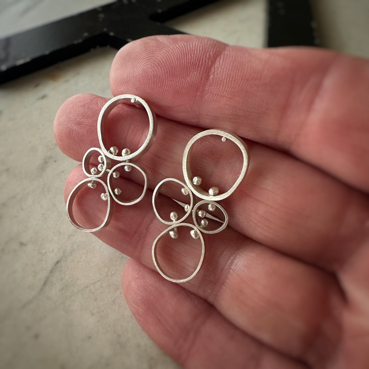 A pair of silver-toned geometric dangle earrings with small bead accents, held in a person's hand against a neutral surface.