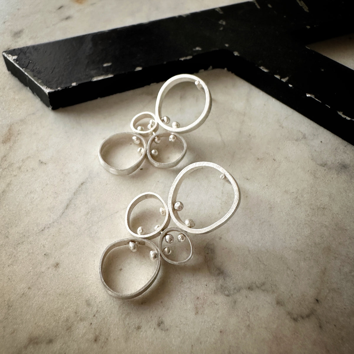 A pair of silver statement earrings with organic, irregularly shaped rings and small bead details, displayed on a light marble surface next to a black wooden block.