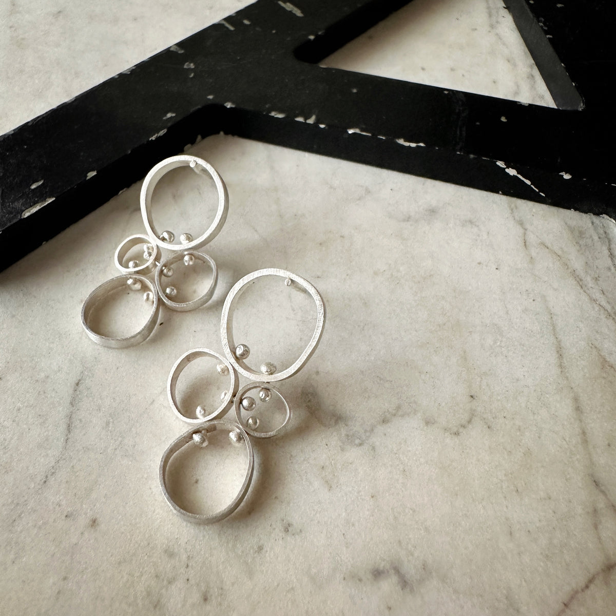 A pair of silver dangle earrings with multiple nested circular rings and small bead details, displayed on a light marble surface next to a black decorative object.