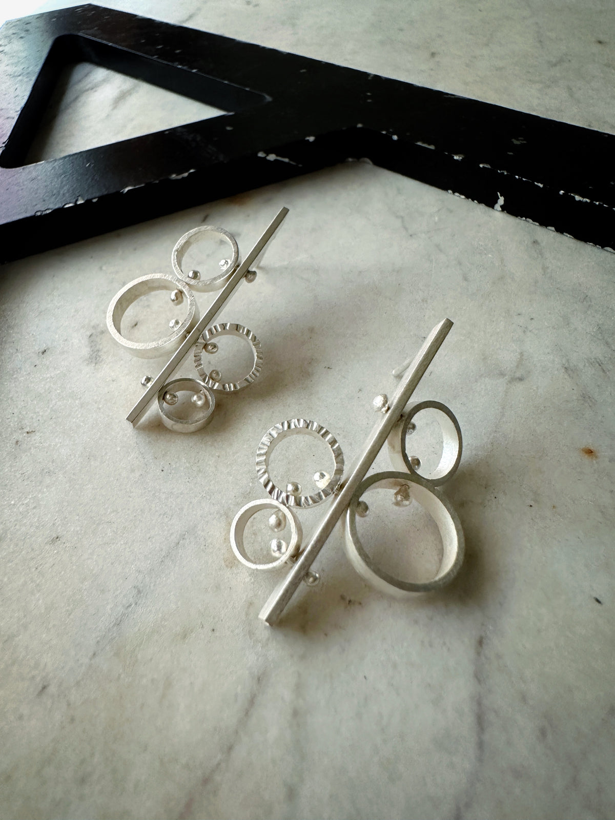 A pair of modern silver geometric earrings with stacked rings and diagonal bars, displayed on a light marble surface next to a black decorative letter 'A'.