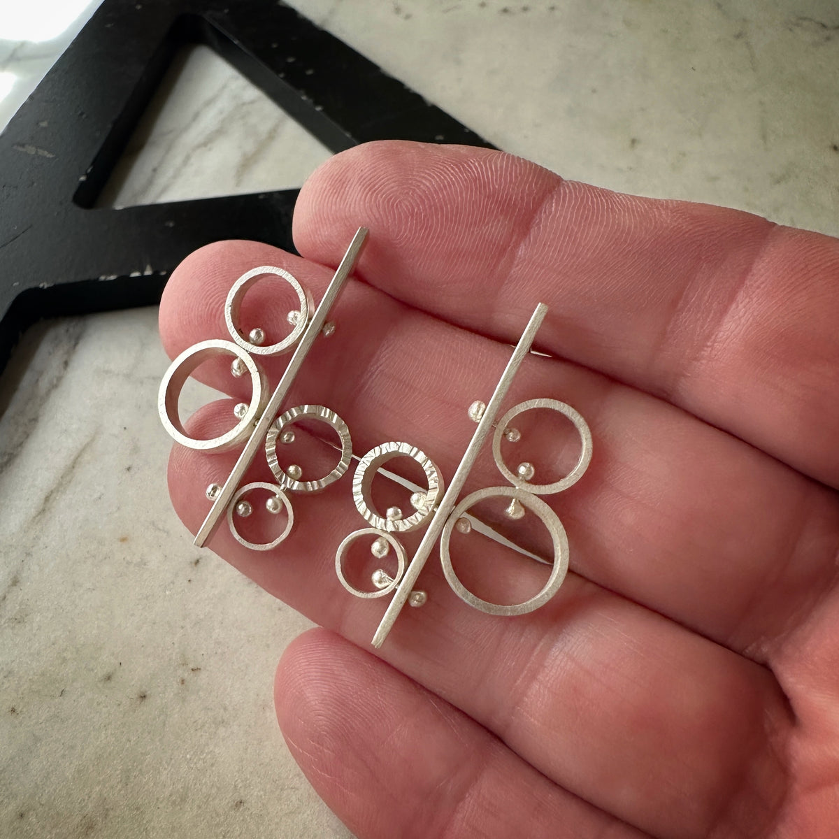 A pair of silver-toned statement earrings with abstract circular and linear designs, held on a person's fingers against a light marble surface.