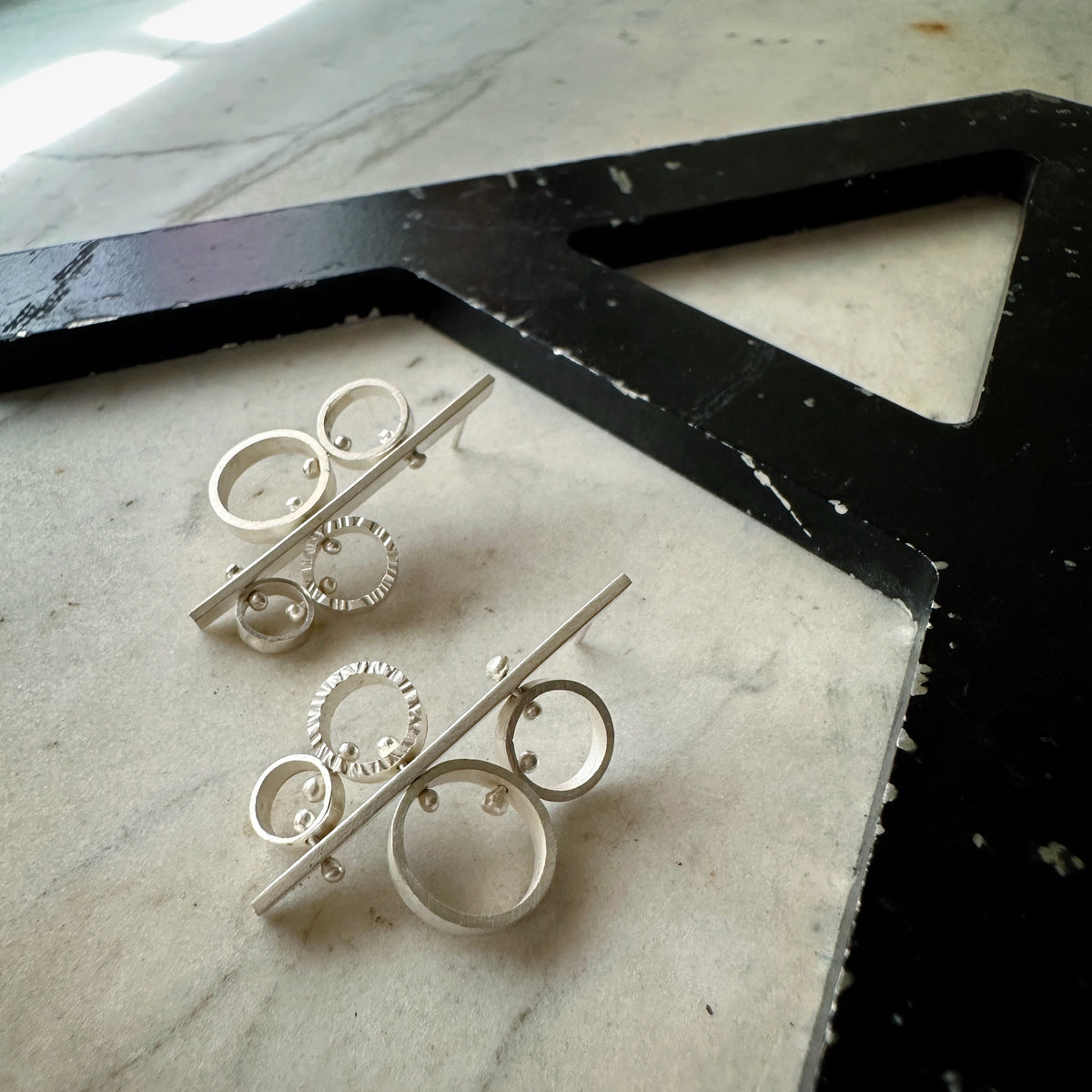 A pair of silver geometric hoop earrings with circular and linear elements, displayed on a light marble surface next to a black decorative frame.