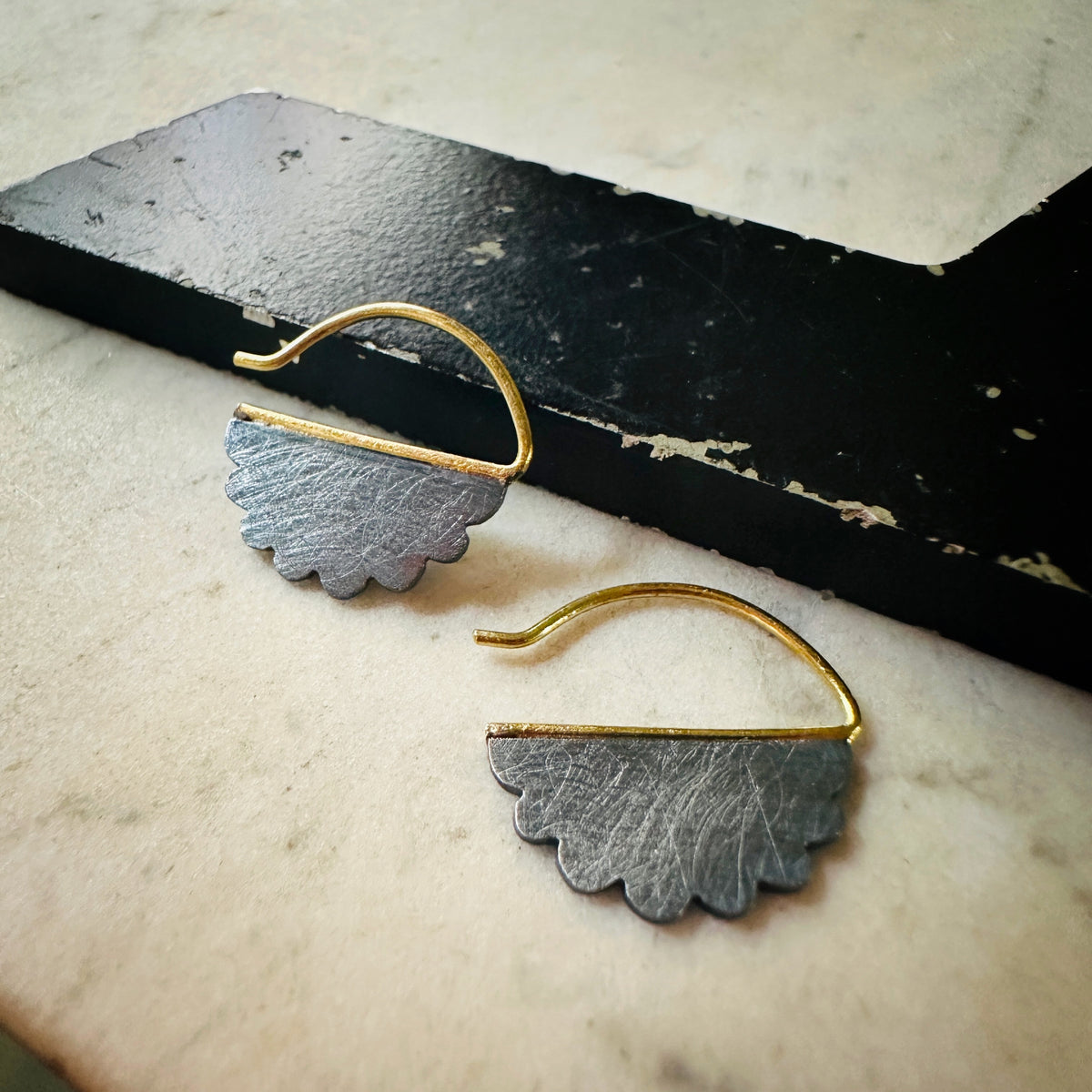 A pair of handmade cloud shaped gold and oxidized silver earrings with curved gold ear wires, displayed on a light marble surface next to a black wooden block.
