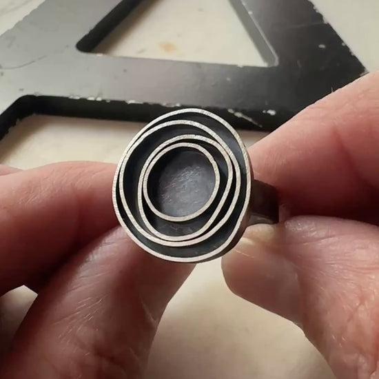 A close-up of a hand holding an oxidized silver ring with concentric circle designs, with a large black metal letter 'A' in the background.