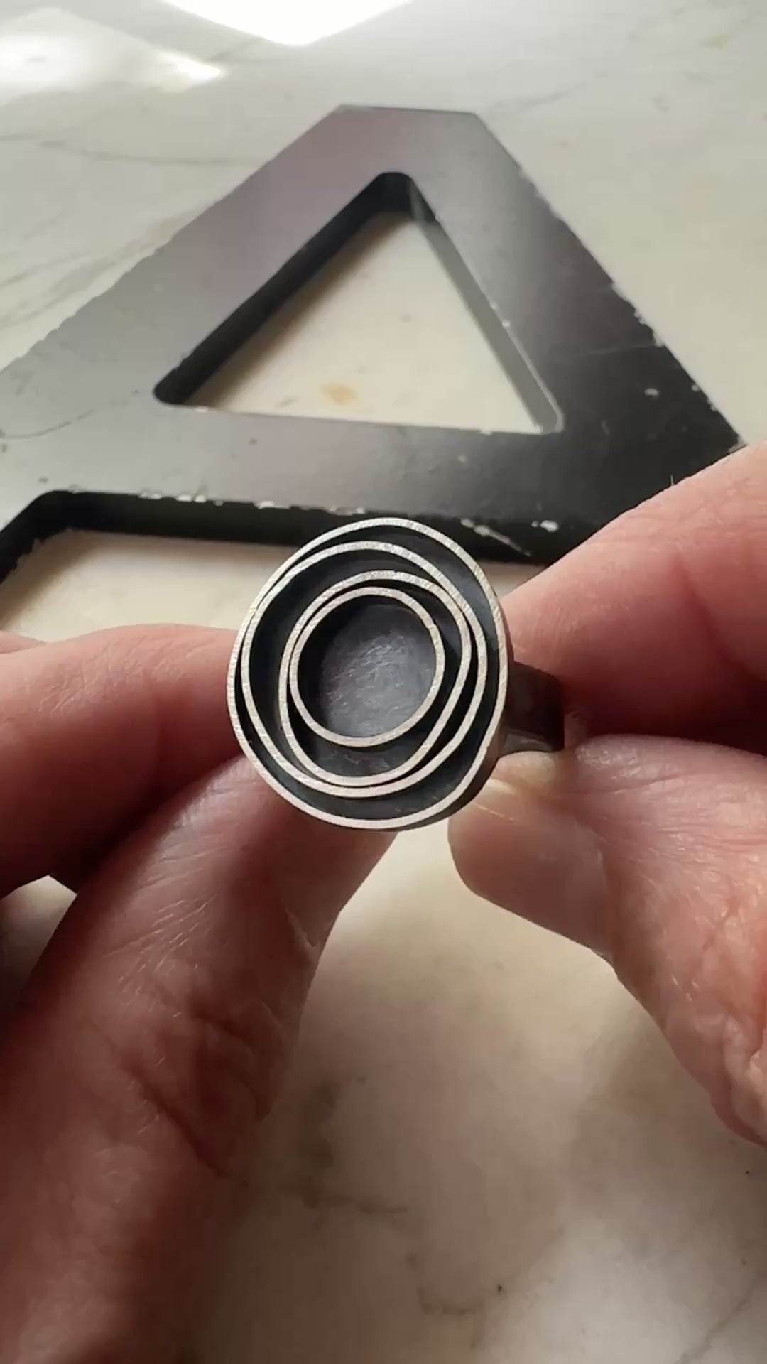 A close-up of a hand holding an oxidized silver ring with concentric circle designs, with a large black metal letter 'A' in the background.