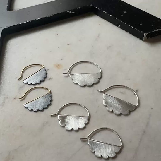 two pair of silver cloud shaped hoop earrings with scalloped edges, displayed on a light beige marble surface next to a black wooden block.