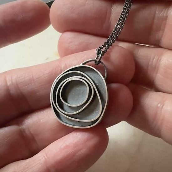An oxidized sterling silver concentric circle pendant necklace on an oxidized silver chain, being held in a hand for scale