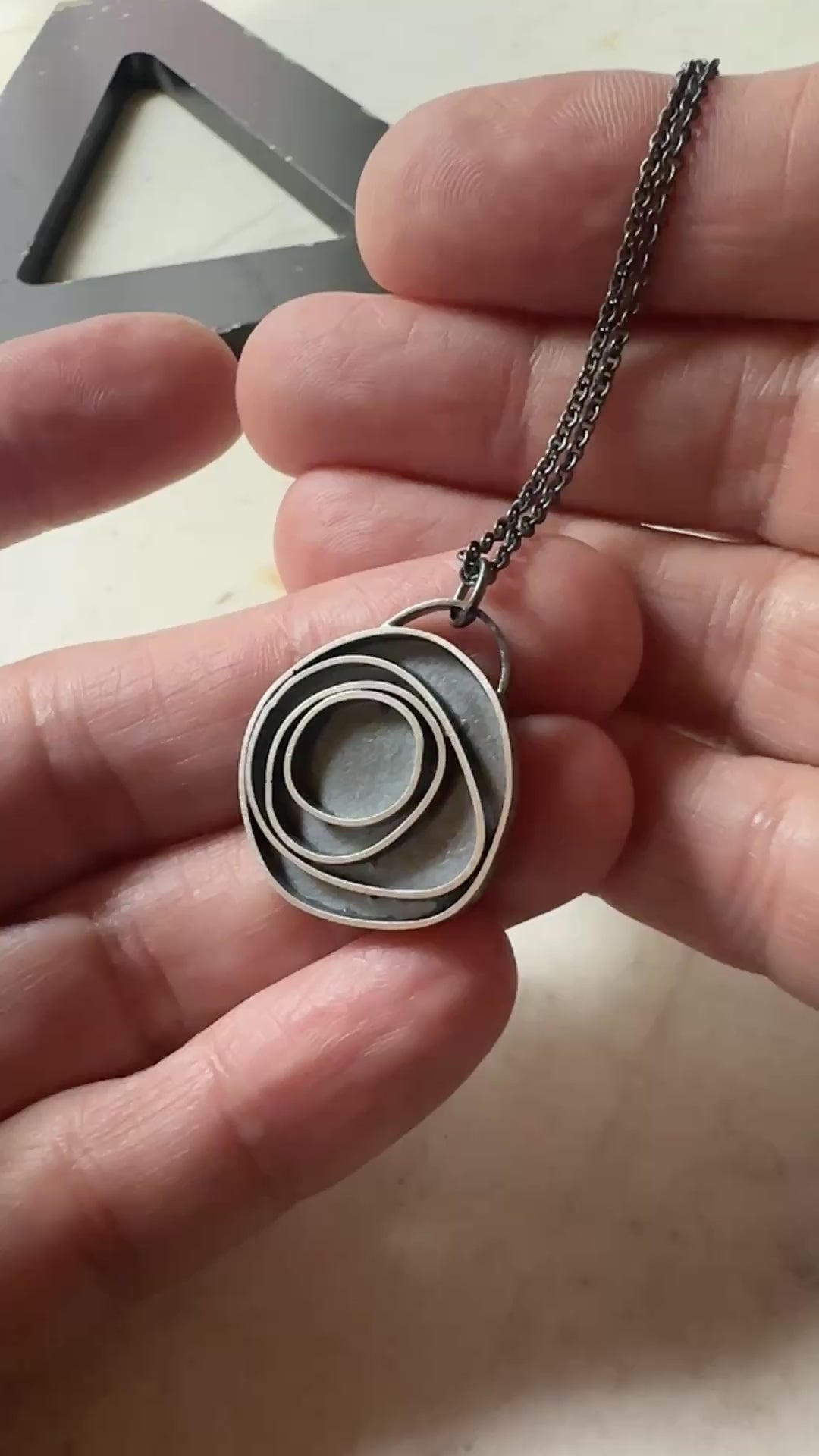 An oxidized sterling silver concentric circle pendant necklace on an oxidized silver chain, being held in a hand for scale