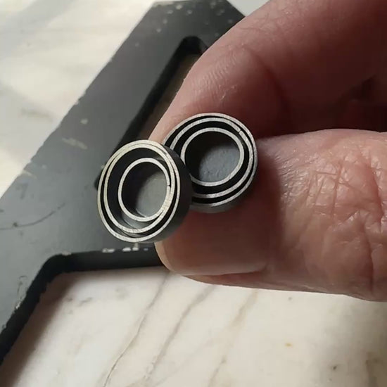 A pair of round stud earrings with a concentric circular design in a oxidized sterling silver, resting on a light beige marble surface.