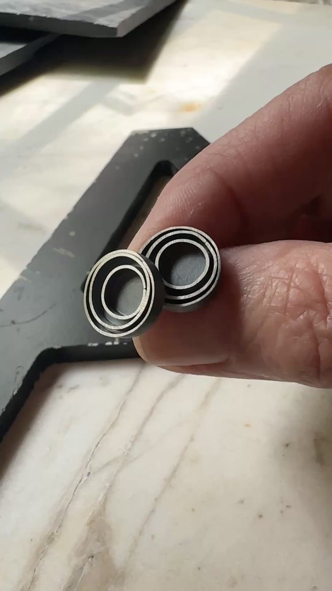 A pair of round stud earrings with a concentric circular design in a oxidized sterling silver, resting on a light beige marble surface.