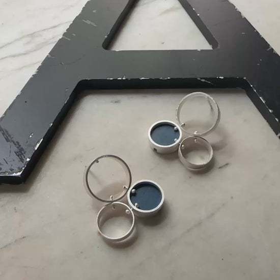 A pair of silver post  earrings with cellular drop elements featuring small round blue formica, displayed on a light marble surface.