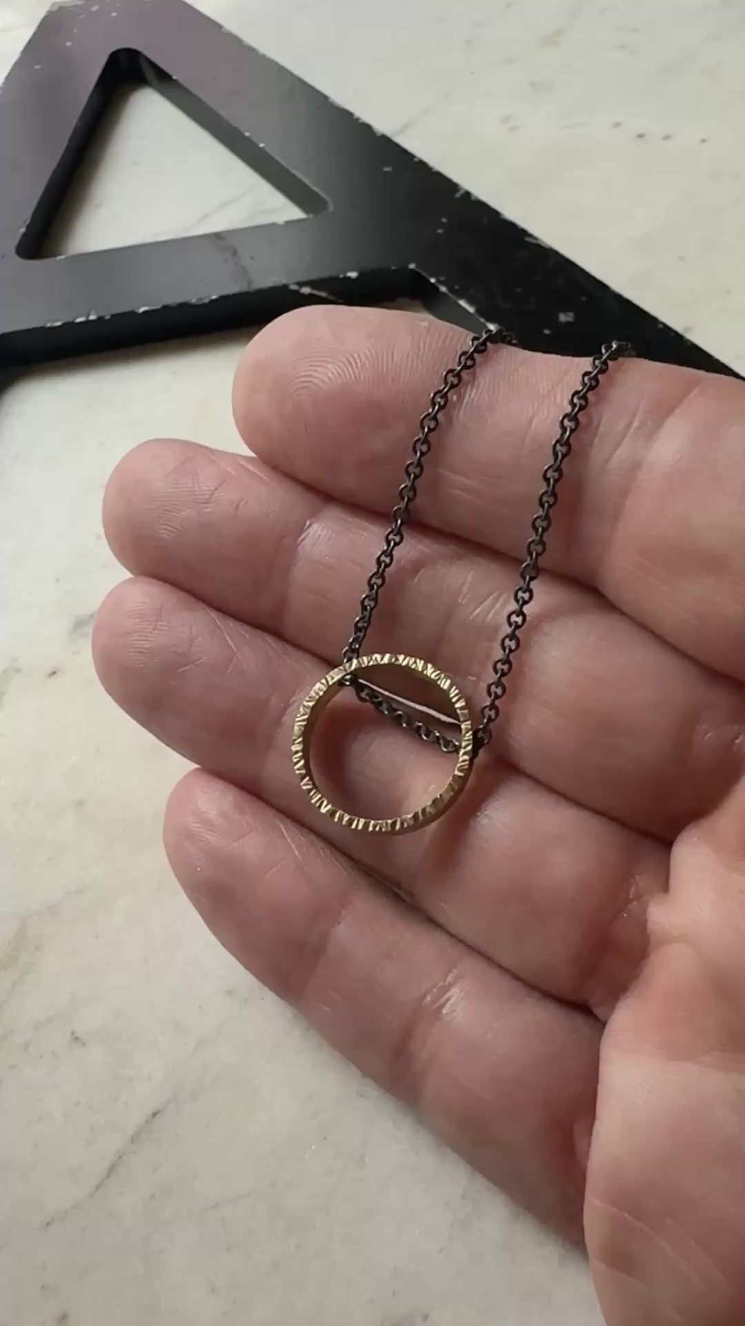 yellow gold hammered texture circle pendant with oxidized sterling silver chain on a marble surface 
