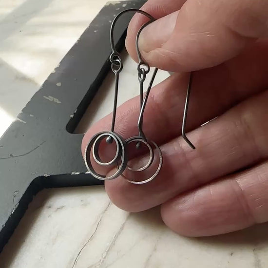 A pair of oxidized sterling silver drop earrings with circular spiral designs, resting on a light beige marble surface next to a black wooden frame.